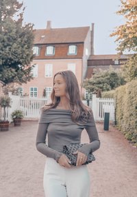 Γκρι μπλούζα με μακριά μανίκια και ανοιχτούς ώμους, λευκά παντελόνια με ψηλή μέση, κρατώντας ένα μαύρο τσάντα με σχέδια, στέκεται σε μια πεζοδρόμιο με χαλίκι και πράσινα φυτά.