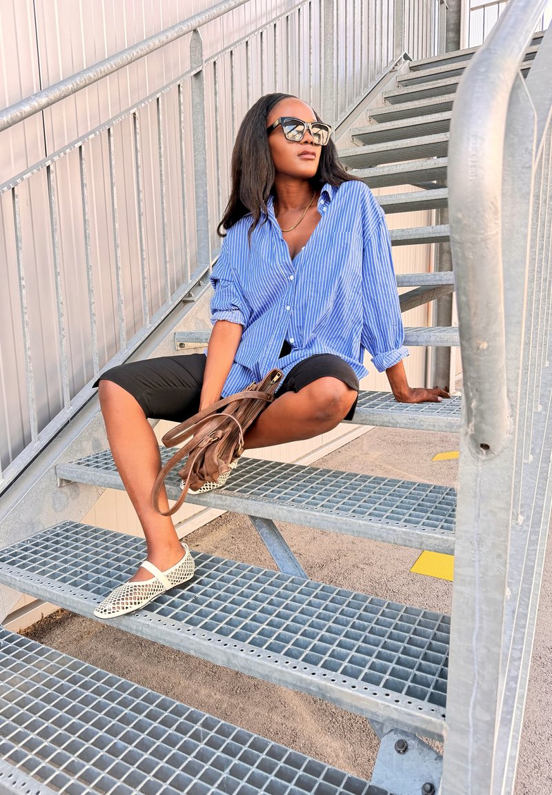 Femme portant des lunettes de soleil, une chemise à rayures bleues et un short noir assise sur des escaliers métalliques extérieurs tenant un sac à main marron.