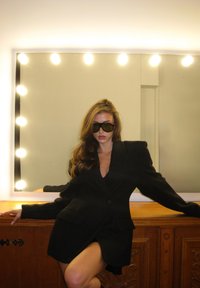 Blazer noir oversize avec épaulettes, décolleté en V profond et deux poches, porté avec des lunettes de soleil foncées. Fond miroir éclairé, surface en bois.