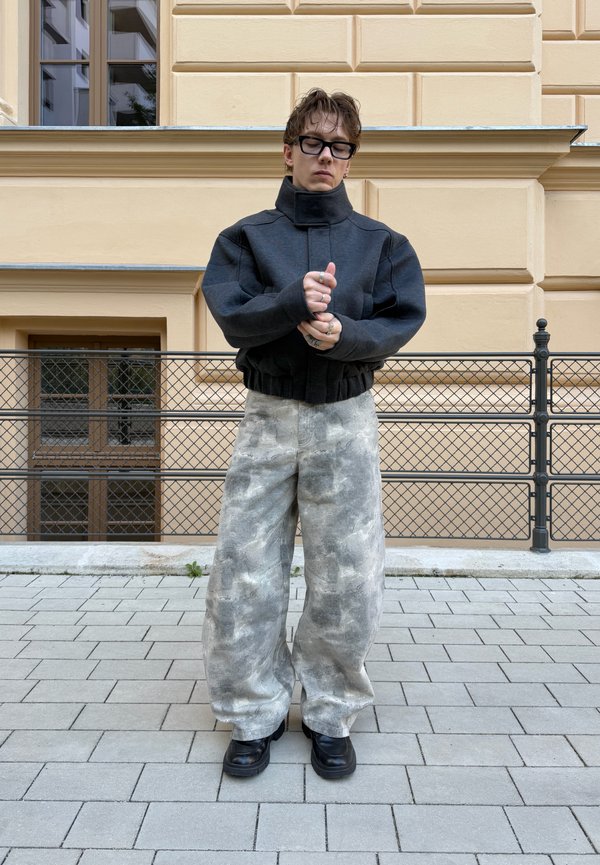 Une personne portant des lunettes, une veste sombre à col montant, un pantalon gris ample à motifs et des chaussures noires se tient devant un bâtiment en pierre de couleur crème.