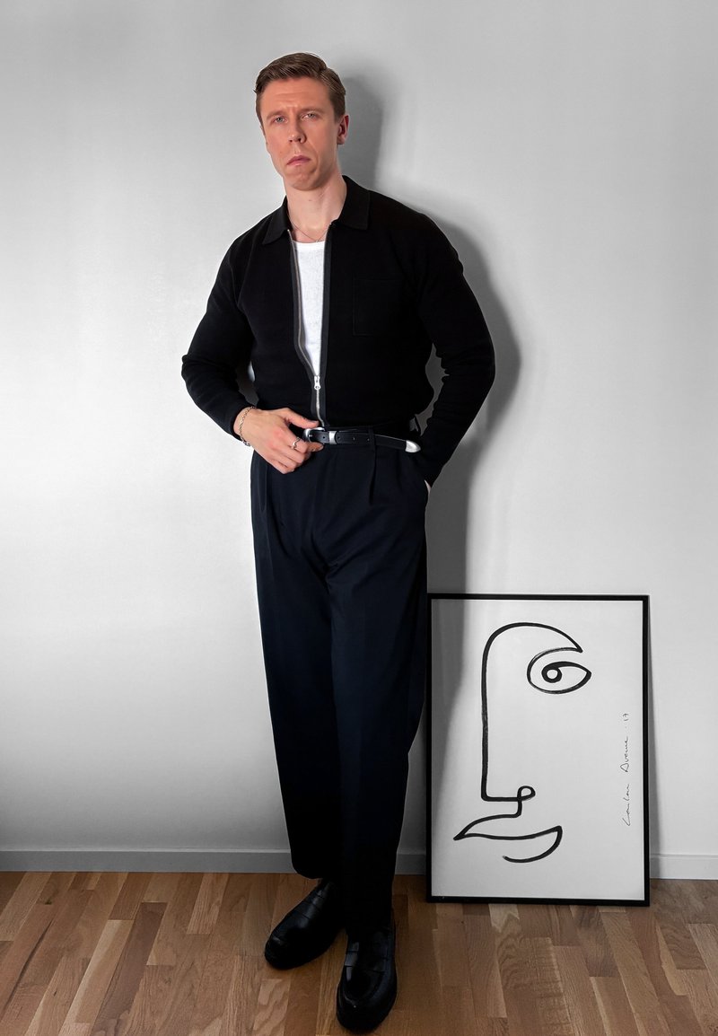 Homme en veste et pantalon noirs, debout à côté d'un dessin minimaliste au trait noir encadré d'un visage sur un sol en bois contre un mur blanc.