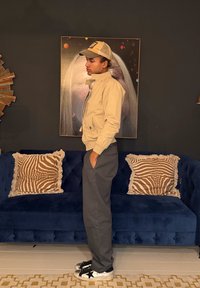 Jeune homme en veste beige et casquette, debout de profil devant un canapé bleu foncé avec des coussins à rayures zébrées et une œuvre murale abstraite.