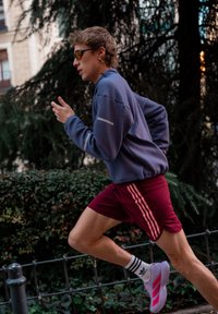Jonge persoon die buiten hardloopt, gekleed in een paarse jas, bordeauxrode short, witte sokken, witte schoenen en sports zonnebril met oranje tint.