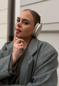 Over-ear koptelefoon in zilver en beige gedragen door een persoon die een grijze wollen jas draagt, met een vinger bedachtzaam op de lippen gelegd.