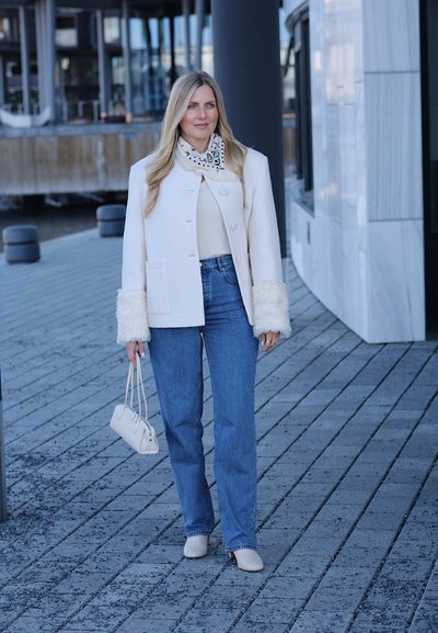 Mujer con chaqueta blanca con puños de piel, jeans azules, botas blancas y bufanda estampada, caminando por una acera urbana pavimentada y llevando un bolso pequeño.