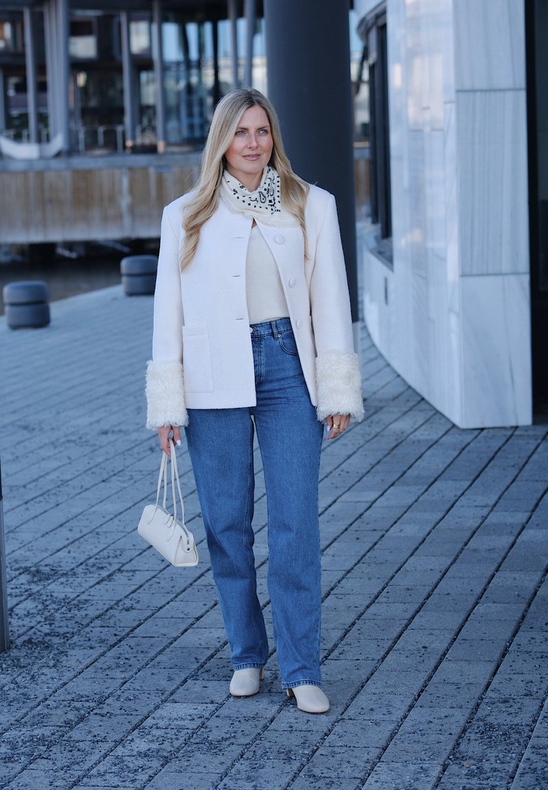Femme portant une veste blanche avec des poignets en fourrure, un jean bleu, des bottes blanches et une écharpe à motifs, marchant sur un trottoir urbain pavé en tenant un petit sac à main.