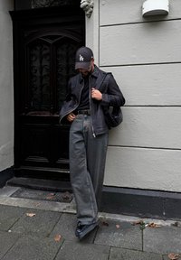 Černá kožená bunda, černé tričko, šedé široké džíny, černá kšiltovka s logem "LA" a černé boty. Stojící vedle zdobených dřevěných dveří.