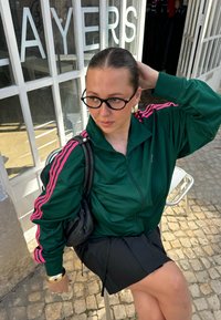 Green zip-up track jacket with pink stripes, black pleated skirt, and glasses. Sitting on a silver chair against a glass shopfront.