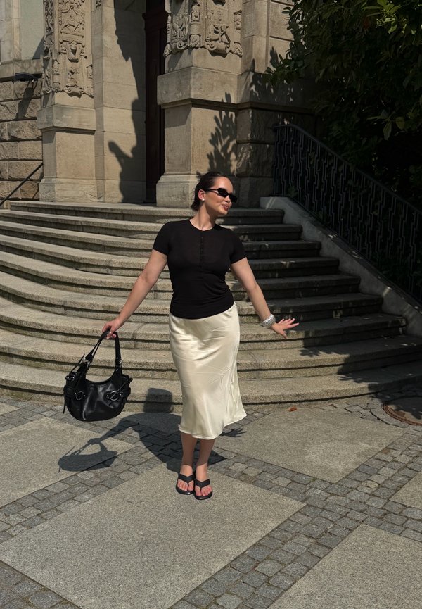 Femme portant des lunettes de soleil, un haut noir et une jupe crème, tenant un sac à main noir, debout près d'escaliers en pierre et d'une façade de bâtiment ornée au soleil.