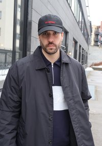 Άνδρας με γένια και σκουλαρίκια, που φοράει μαύρο καπέλο και σκούρο μπουφάν, στέκεται έξω σε αστική περιοχή με μοντέρνα κτίρια στο φόντο.