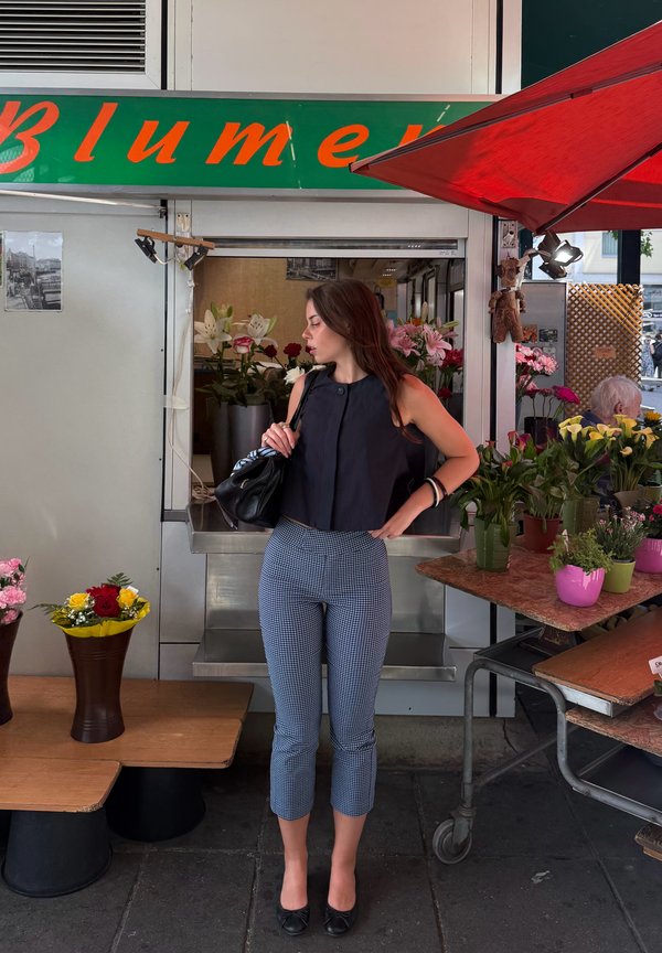 Woman in black top and patterned pants stands near flower shop window with bouquets and plants on display tables.