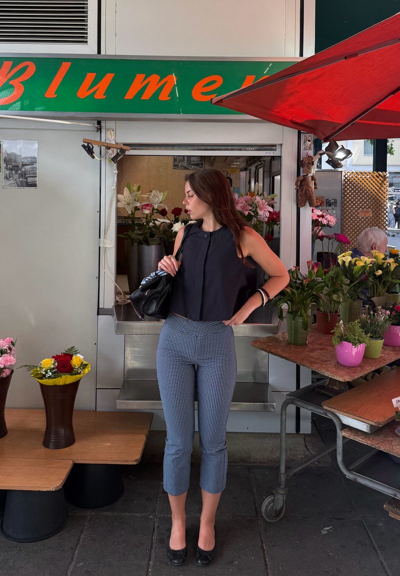 Femme en haut noir et pantalon à motifs se tient près de la vitrine d'une boutique de fleurs avec des bouquets et des plantes sur des tables d'exposition.