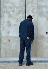 Giubbotto di denim e pantaloni a righe sottili di colore blu scuro. Il giubbotto presenta un colletto alto e un'etichetta sul retro. Tessuto testurizzato con cuciture visibili.