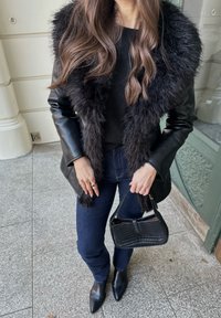 Črna usnjena jakna z ovratnikom iz umetnega krzna, kombinirana s črno majico in temnimi kavbojkami iz jeansa. Dodatki vključujejo teksturirano črno torbico in konične škornje.