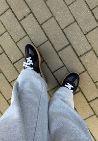 Μαύρα και γκρι αθλητικά παπούτσια με άσπρα κορδόνια, διαθέτοντας ένα μείγμα υλικών και σόλα από καουτσούκ, εικονιζόμενα από πάνω με γκρίζα παντελόνια.