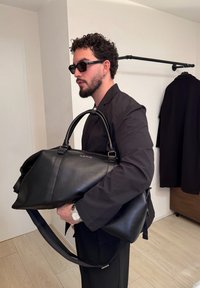 Homme aux cheveux bouclés et à la barbe, portant des lunettes de soleil et une veste noire, tenant un grand sac en cuir noir Valentino dans une pièce minimaliste.