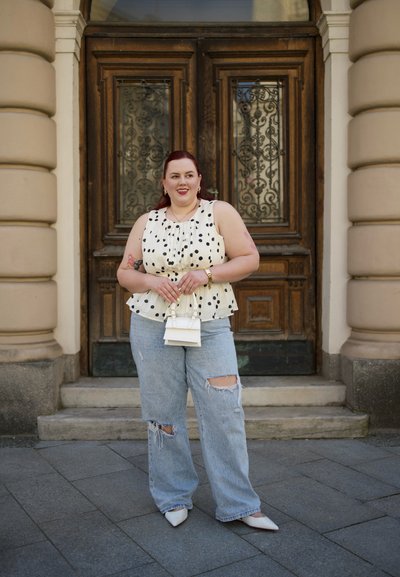 Blusa peplum blanca con puntos negros, jeans rotos azul claro y tacones blancos de punta. Sosteniendo un pequeño bolso blanco.