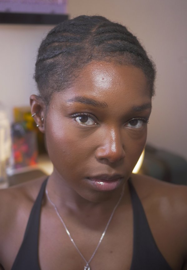 Vrouw met gevlochten haar en zilveren oorbellen, gekleed in een zwarte top en een zilveren ketting, kijkt recht in de camera binnen.