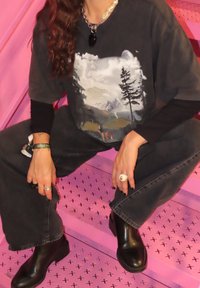 Čierne tričko s grafickým motívom hôr, v kombinácii s čiernymi džínsami s širokými nohavicami a členkovými čižmami. Doplnky zahŕňajú prstene a výrazný náhrdelník.