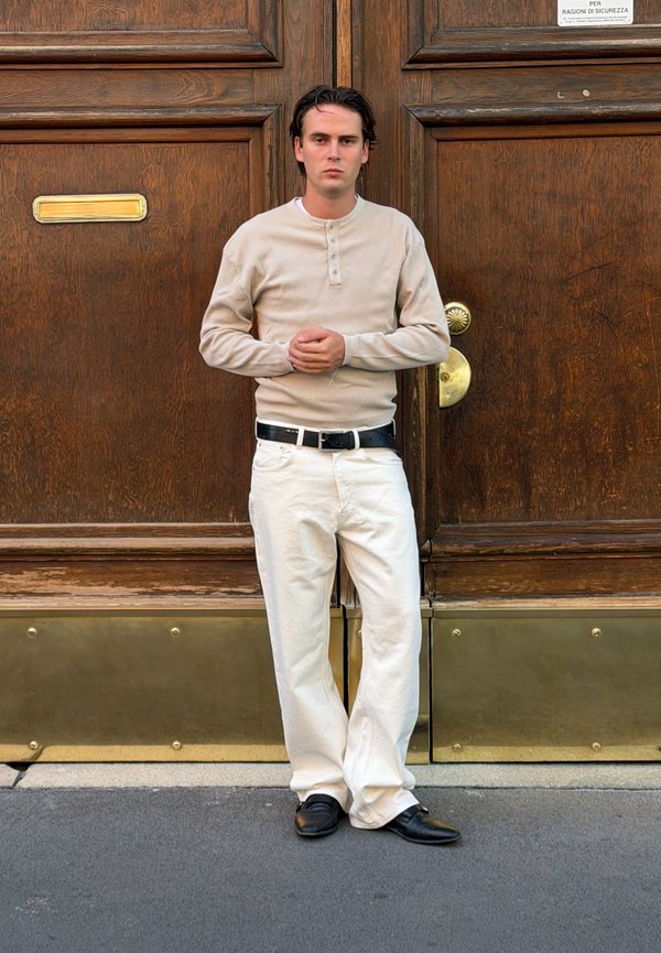 Joven con camisa beige de manga larga y pantalones blancos de pie con las manos entrelazadas frente a grandes puertas de madera con detalles en latón.