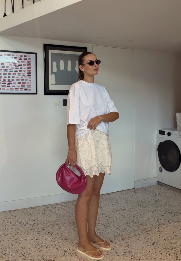 Vrouw draagt wit oversized overhemd, kanten rok, slippers, zonnebril, houdt een roze handtas vast, staat binnen nabij een wasmachine en ingelijste muurkunst.