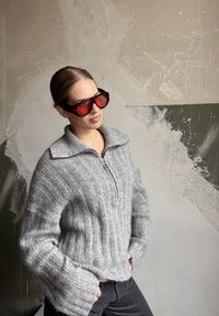 Grey ribbed knit jumper with a zip-up collar and wide sleeves, paired with large black sunglasses with red lenses.