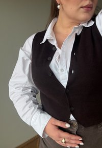 Dark brown, fitted waistcoat over a white button-up shirt. The waistcoat features six black buttons and a textured finish. Model's hands on hips.