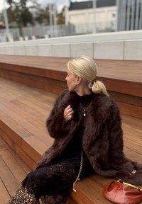 Smeđa umjetna krzna kaput preko crne čipkaste haljine, ukrašena slojevitim zlatnim ogrlicama i sunčanim naočalama, sjedi na drvenim stepenicama. Narančasta torba u blizini.