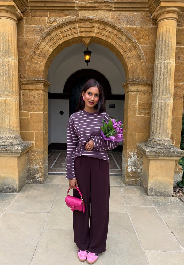 Une femme tenant des tulipes violettes et un sac à main rose vif se tient sous une arche en pierre. Elle porte un haut rayé, un pantalon foncé et des chaussures roses.