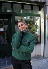 Sudadera de forro polar verde con cuello alto, bolsillo frontal y botones azul marino. Combinada con pantalones negros, sobre un fondo lleno de plantas.