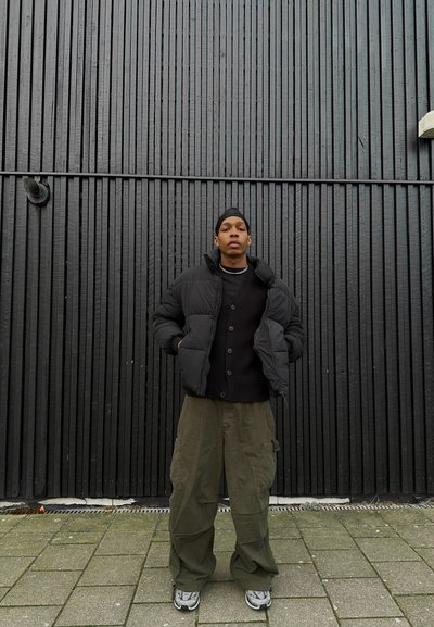 Chaqueta acolchada negra con cremallera y capucha, superpuesta a un cárdigan negro. Pantalones cargo verde oliva y zapatillas grises completan el atuendo.