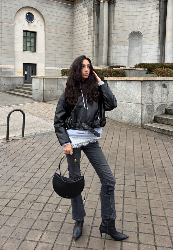 Femme aux longs cheveux noirs portant une veste en cuir noire, un jean gris et des bottes noires, tenant un sac à main noir, debout sur une surface pavée en pierre.