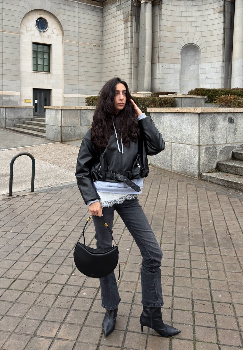 Femme aux longs cheveux noirs portant une veste en cuir noire, un jean gris et des bottes noires, tenant un sac à main noir, debout sur une surface pavée en pierre.