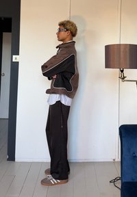 Άτομο με σγουρά μαλλιά που φοράει γυαλιά ηλίου, καφέ και μαύρο μπουφάν, ριγέ πουκάμισο, μαύρο παντελόνι και γκρι αθλητικά παπούτσια, στέκεται με τα χέρια σταυρωμένα σε εσωτερικό χώρο.