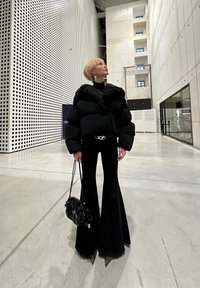 Pessoa com cabelo loiro curto, vestindo uma jaqueta puffer preta e calças largas, segurando uma bolsa preta, de pé em um edifício moderno e espaçoso.