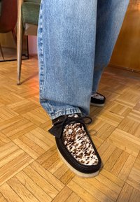 Personne portant un jean bleu et des chaussures à lacets noires avec un détail de fourrure tachetée marron et blanche, debout sur un parquet en bois.