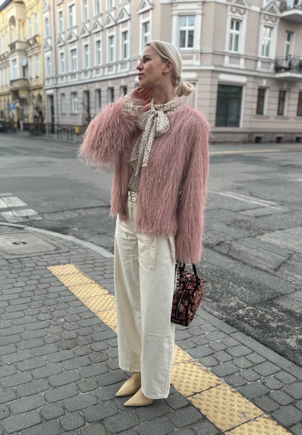 Vrouw met blond haar in een roze pluizige jas, crèmekleurige sjaal met stippen, wijde witte broek, beige laarzen, met een patroon op de tas, op een stadsstoep.