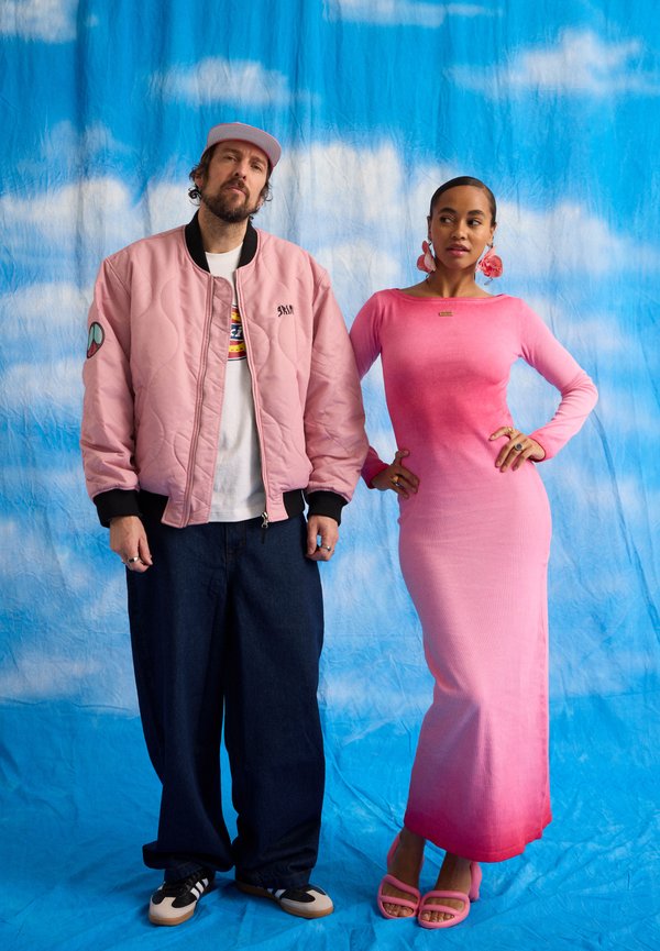 Hombre con chaqueta bomber rosa, gorra, pantalones negros y zapatillas de deporte, de pie junto a mujer con vestido ombré rosa y tacones rosas a juego, con un cielo azul de fondo.