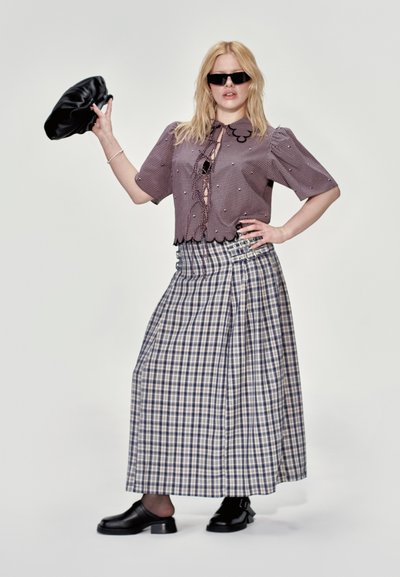 Blusa de manga corta a cuadros en púrpura y blanco con adornos decorativos, combinada con una falda larga a cuadros y mocasines negros. La modelo sostiene un bolso negro.