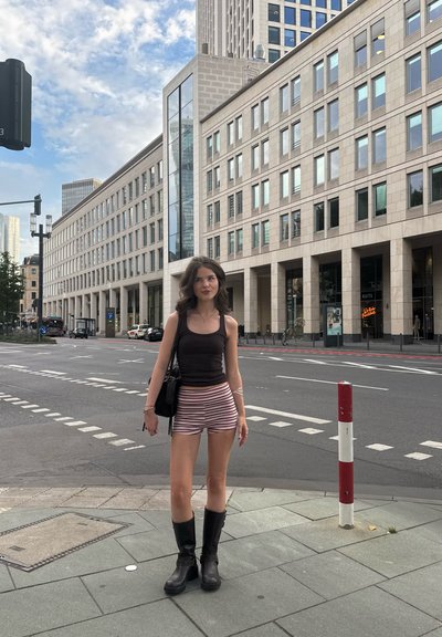 Top negro sin mangas, pantalones cortos a rayas y botas negras hasta la rodilla. De pie en una calle de la ciudad con edificios modernos y marcas viales visibles.