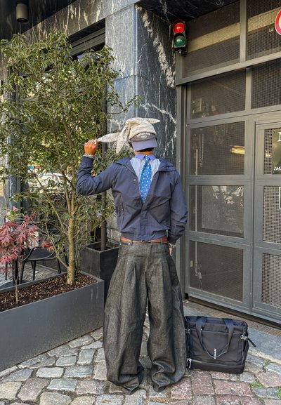 Persona con pantalones oscuros de gran tamaño, chaqueta azul, corbata azul, gorra gris, sosteniendo un suéter gris, de pie cerca de macetas y una puerta metálica gris.