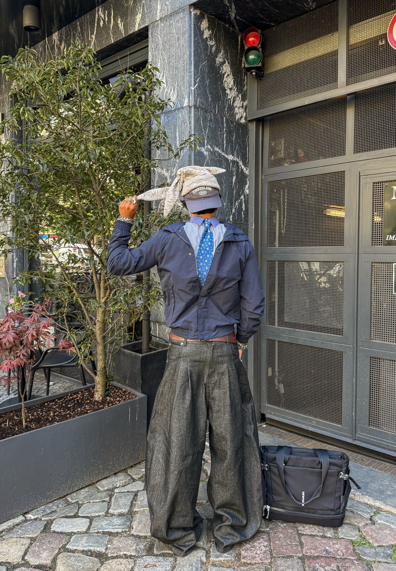 Personne portant un pantalon sombre surdimensionné, une veste bleue, une cravate bleue, une casquette grise, tenant un pull gris, debout près de plantes en pot et d'une porte métallique grise.