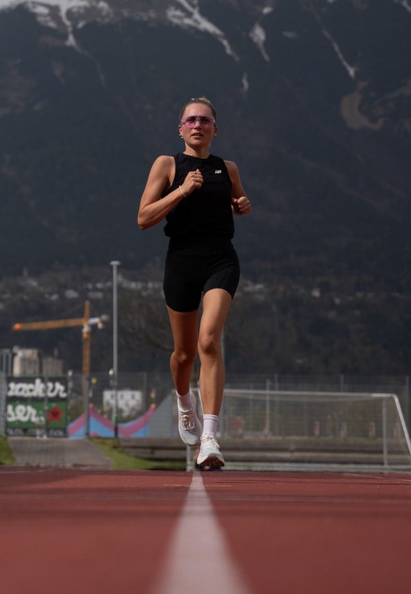 Schwarzes sportliches Tanktop und Shorts, weiße Laufschuhe. Läufer auf einer roten Laufbahn mit Bergen im Hintergrund und einer verschwommenen Baustelle.
