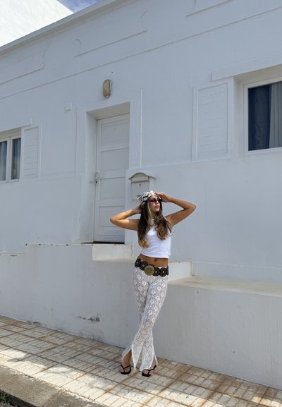Top corto blanco, pantalones de encaje blancos y un cinturón negro con acentos dorados. El fondo presenta un edificio blanco con una puerta y una ventana.