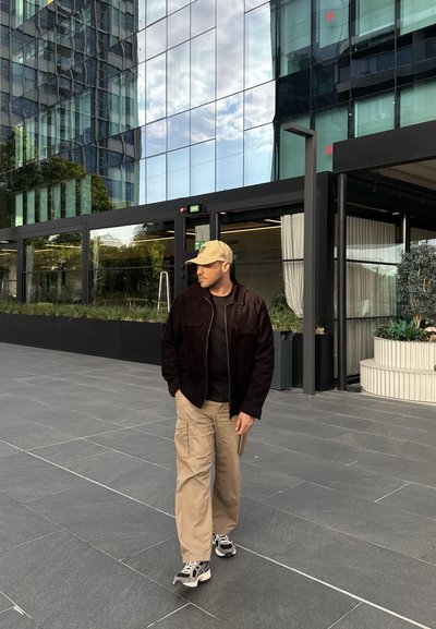 Chaqueta de pana oscura, camiseta negra, pantalones cargo caqui y zapatillas deportivas. De pie sobre una superficie de baldosas grises con un fondo de un edificio moderno de vidrio.