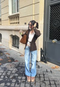 Žena sa tamnom kosom i naočalama drži ledeni napitak, nosi smeđi kardigan, bijelu majicu, svijetlo plave traperice i smeđe cipele, stojeći na gradskom pločniku.