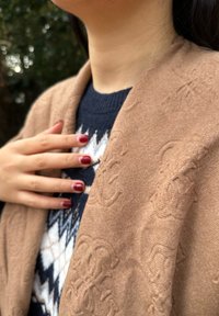 Xale castanho com padrão, colocado sobre um suéter azul-marinho, apresentando um desenho geométrico. Dedos longos com unhas vermelhas repousam suavemente sobre o tecido.