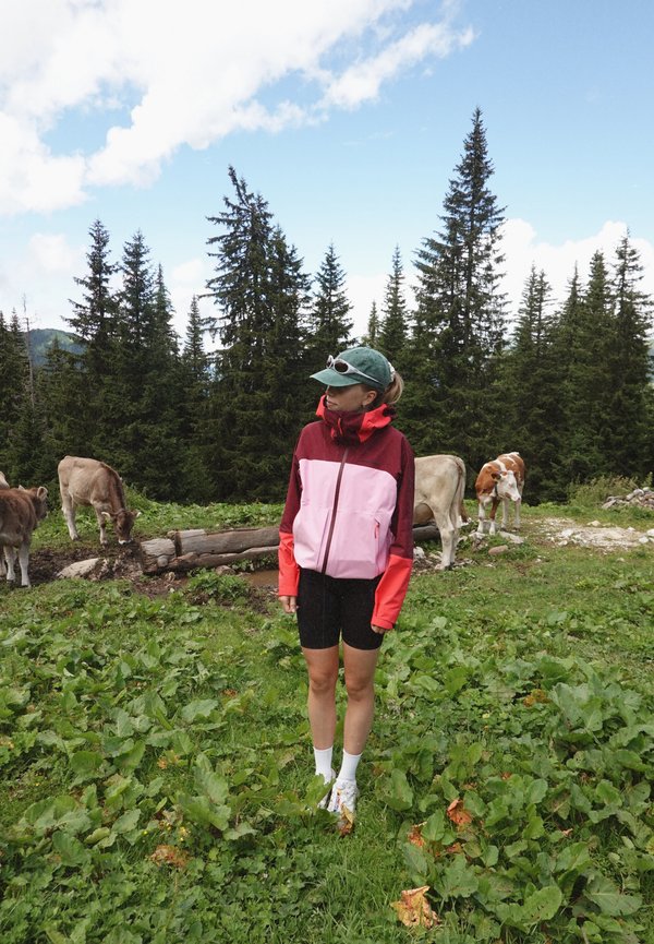Červená, ružová a oranžová nepremokavá bunda s vysokým golierom a zipsom, skombinovaná s čiernymi šortkami. Zelená čiapka a biele ponožky viditeľné. V pozadí kravy.