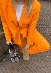 Costume orange sur mesure avec taille ceinturée et pantalon large, associé à un haut blanc, des bottines marron et un sac à main à motifs.
