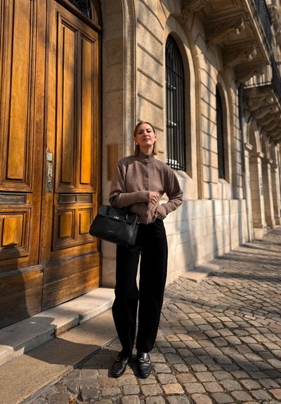 Cárdigan de punto marrón con botones, pantalones de pierna ancha negros y bolso negro. Mocasines brillantes descansan sobre adoquines; puerta de madera al fondo.